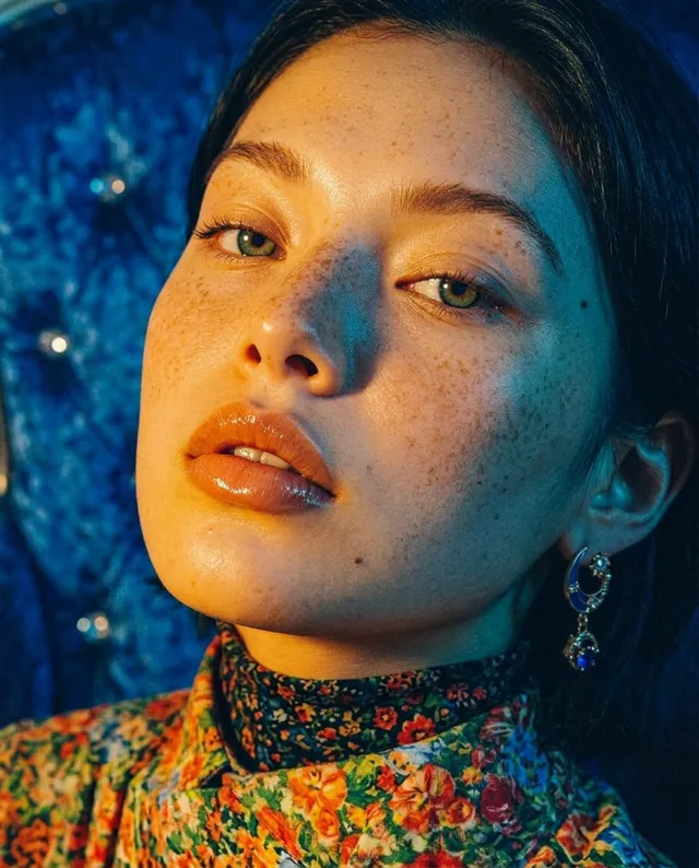 A close-up portrait of a woman with freckled skin and glossy lips, styled in a vibrant floral outfit, exudes a moody, editorial vibe. The image features striking blue and warm tones, creating a contrasting yet harmonious atmosphere.