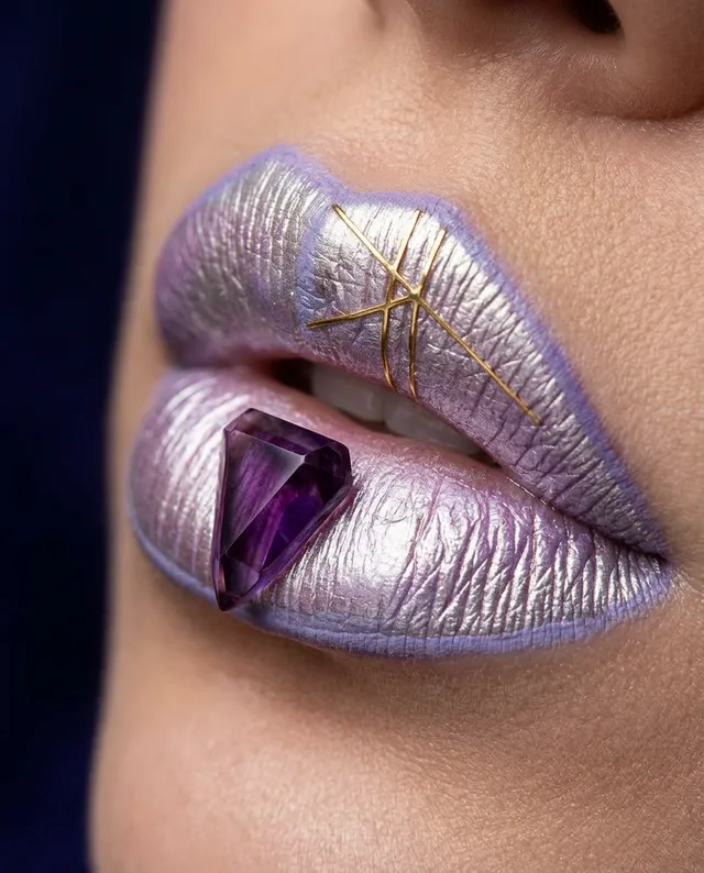 This editorial photography piece features a close-up of metallic lavender and silver lips adorned with a geometric purple gem and delicate golden accents. The mood is avant-garde and surreal, emphasizing a bold and artistic expression.