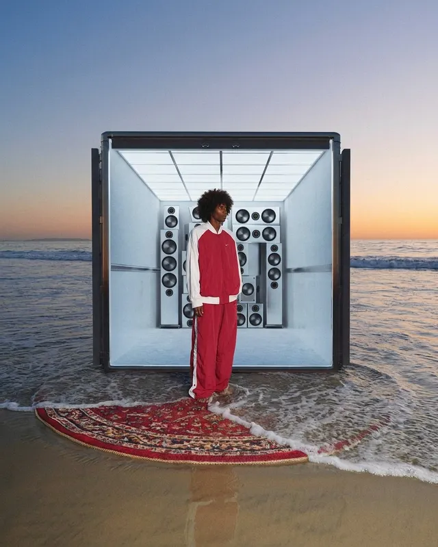 A young person in a striking red tracksuit stands on a rug at the shore, framed by a large sound system emerging from a futuristic, open container. The image blends editorial photography with surreal elements, creating an energetic and vibrant mood, accentuated by the warm tones of the sunset and the cool blues of the water.