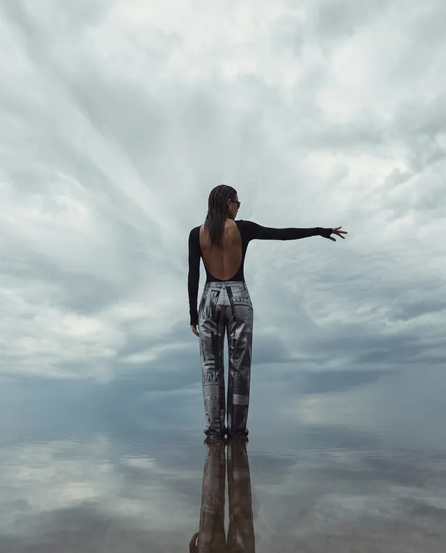 A striking editorial photograph captures a figure in a sleek black backless top and patterned pants, standing in a reflective expanse under moody, swirling gray clouds. The somber color palette and dramatic atmosphere evoke a sense of introspection and solitude.