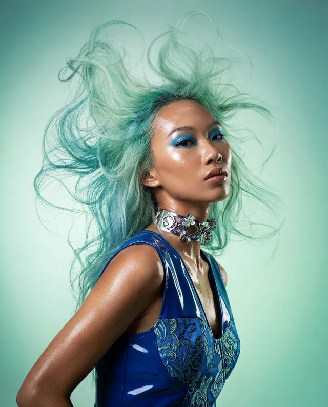 A striking editorial photograph features a model with vibrant, flowing aqua hair and bold blue makeup, exuding a confident and dynamic energy. The image is set against a soft green background, enhancing the cool color palette and the model's glossy, futuristic look.