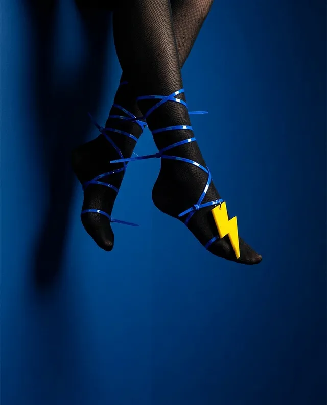 This editorial photography features a striking close-up of feet adorned in black stockings, wrapped in vibrant blue ribbons and highlighted by a bold yellow lightning bolt accessory. The composition is set against a deep blue background, evoking a playful yet energetic mood.