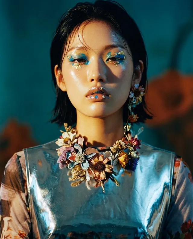This striking editorial photograph features a model adorned with a floral necklace and shimmering makeup, evoking a surreal and ethereal mood. Dominated by shades of blue and metallic silver, the composition captures a harmonious blend of nature and high fashion.
