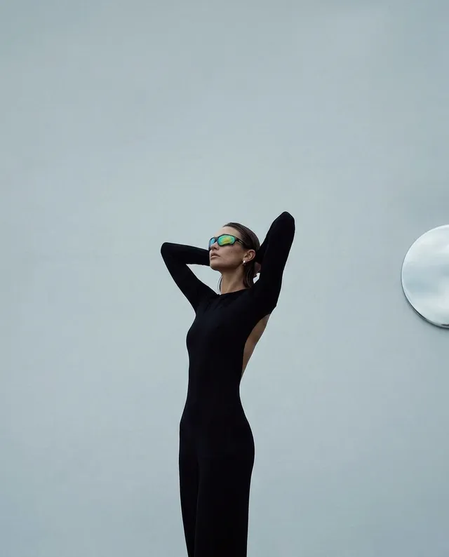 A sleek editorial photograph features a model in a form-fitting black jumpsuit, striking a confident pose against a soft gray wall. The cool tones and minimalistic backdrop convey a modern and sophisticated mood, accentuated by her reflective sunglasses.