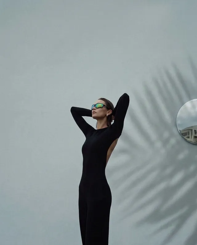 A stylish model poses gracefully in a sleek black outfit, exuding confidence against a minimalist backdrop of soft gray. The editorial photography captures a modern, sophisticated mood enhanced by the shadows and reflections in the environment.
