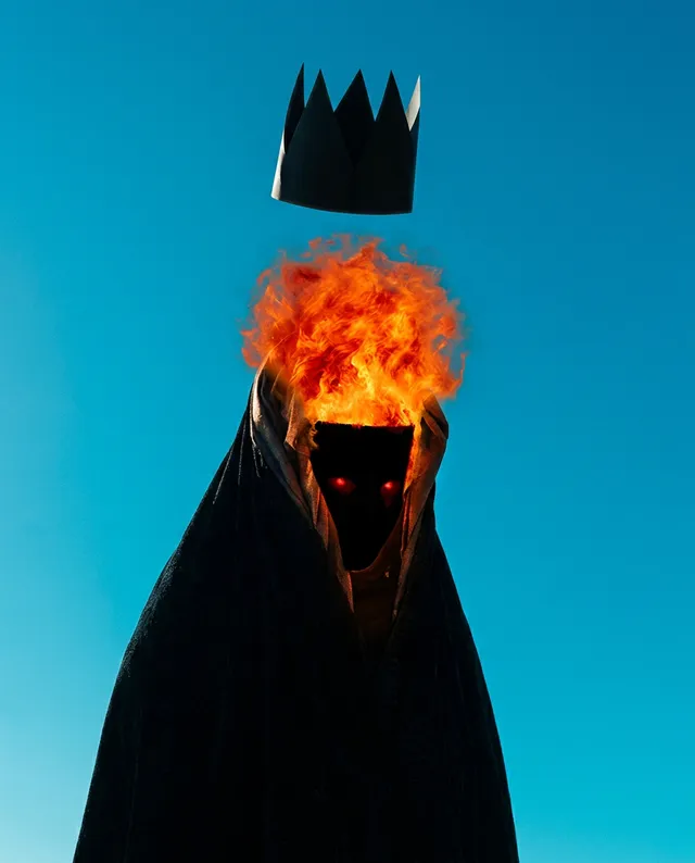 A striking editorial photograph features a cloaked figure with a crown floating above, enveloped in vibrant orange flames that emerge from its head against a crisp blue sky. The surreal composition evokes a mood of power and mystique, contrasting dark and fiery tones.