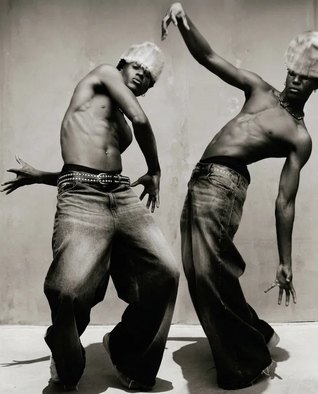 This striking black-and-white editorial photograph features two male models in dynamic poses, showcasing their toned physiques and oversized denim trousers. The mood is edgy and unconventional, highlighted by the stark contrast of light and shadow.