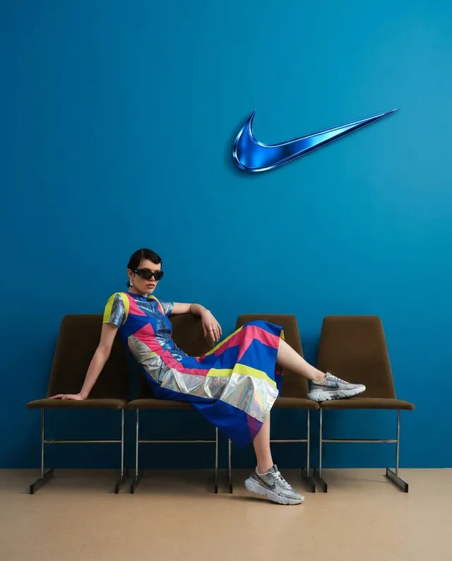 A stylish model poses in a vibrant, patterned dress and sunglasses against a bold blue wall, with the iconic Nike logo overhead. The editorial photography captures a modern, urban mood, featuring a playful contrast of colors.