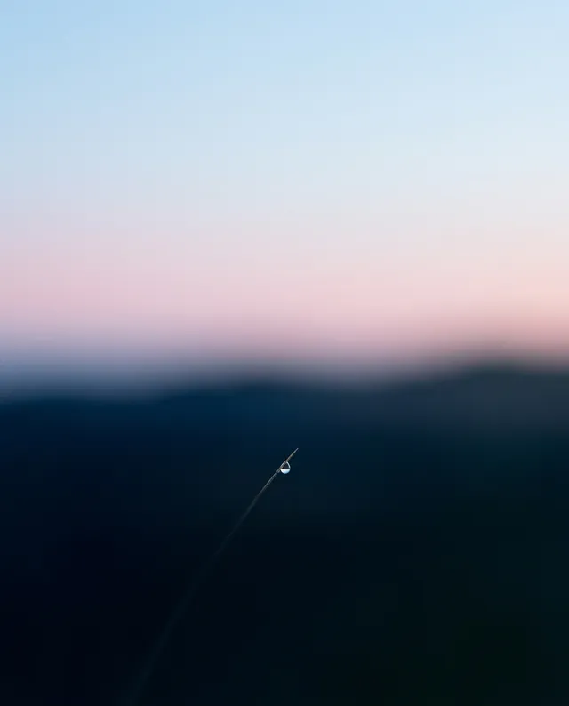 A close-up photograph captures a delicate water droplet resting on a blade of grass, set against a softly blurred pastel background. The mood is serene, with dominant cool colors of blue and pink creating a tranquil atmosphere.