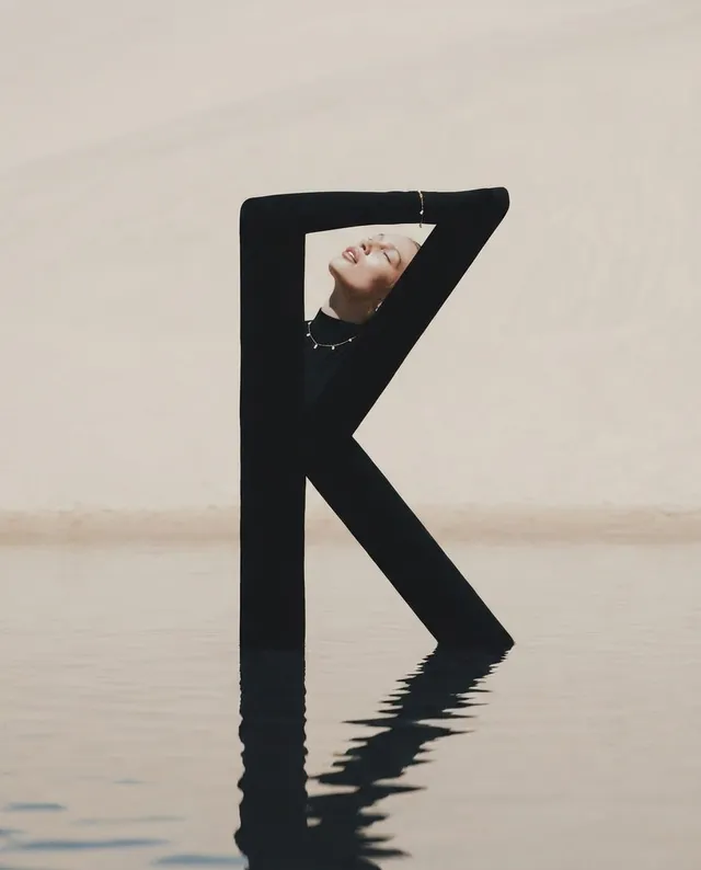 A striking editorial photograph features a model gracefully posed within a large, black letter "K" rising from tranquil water, set against a serene sandy backdrop. The composition exudes a minimalist mood with muted color tones, emphasizing elegance and artistic flair.