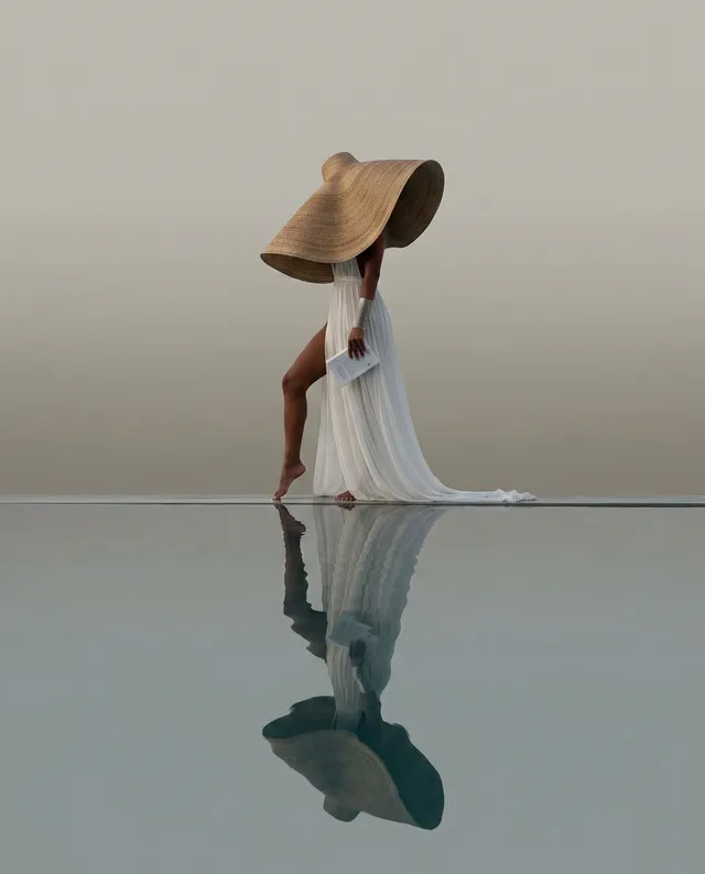 A serene editorial photograph captures a figure in a flowing white dress and an oversized straw hat, standing gracefully by a reflective surface that doubles the serene mood. Dominant soft tones of beige and white enhance the tranquil atmosphere.