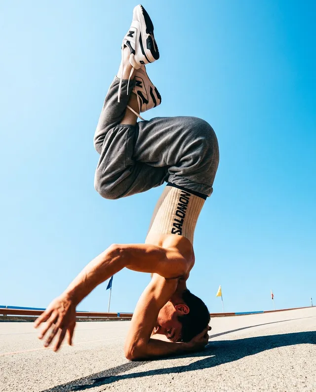 A dynamic editorial photograph capturing a male dancer in a gravity-defying headstand against a clear blue sky. The strong athletic pose is highlighted by a vibrant color palette of blues and grays, evoking a sense of energy and motion.