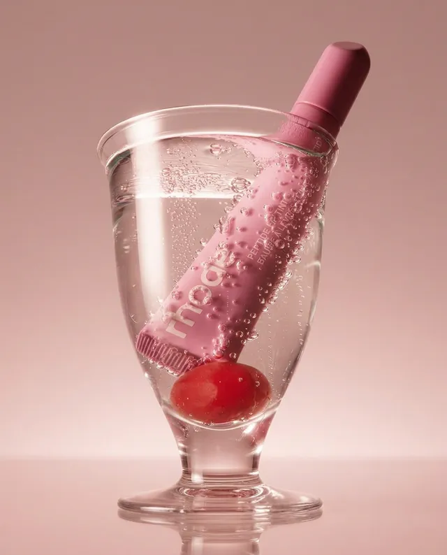 A vibrant pink skincare product stands out in a glass filled with sparkling water, accompanied by a cherry, against a soft pink backdrop. The editorial photography exudes a fresh and playful mood, highlighting a harmonious blend of pastel hues.