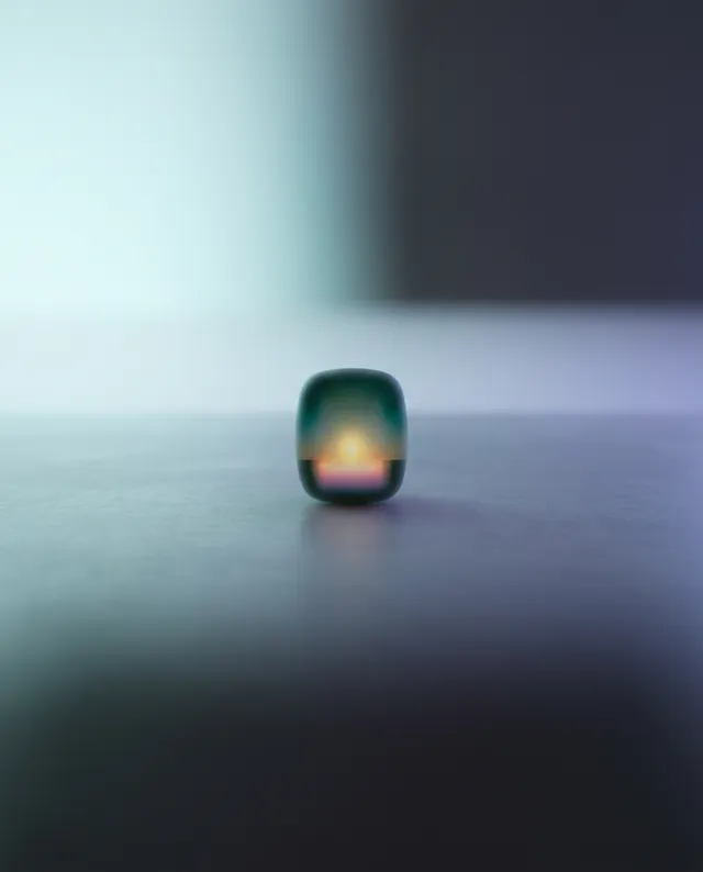 A minimalist close-up of a smooth, rounded object set against a softly blurred background, evoking a tranquil and contemplative mood. The dominant colors are subtle shades of green and gray, with a warm glow emanating from the center of the object, highlighting its shape.