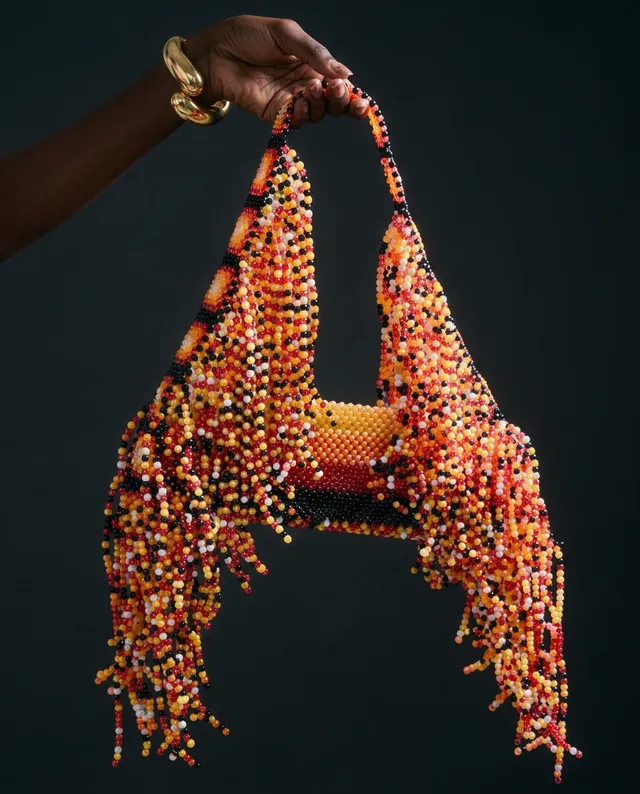 This striking editorial photograph showcases a vibrant, beaded handbag adorned with cascading fringe in warm hues of orange, yellow, and black. The dramatic contrast against a dark background emphasizes the intricate craftsmanship and bold design, evoking a sense of luxury and creativity.