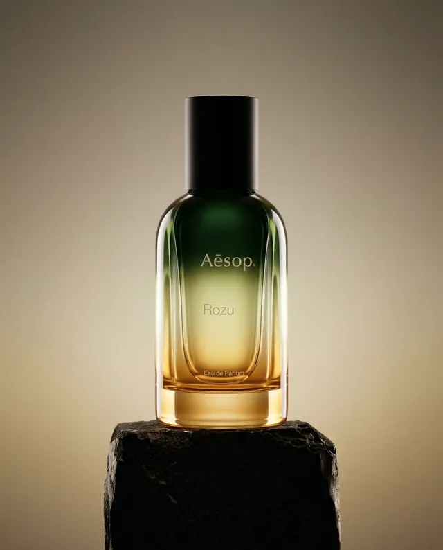 An elegant still life photograph showcasing a bottle of Aesop's Rózu Eau de Parfum, featuring a gradient glass design that transitions from dark green to amber. The image conveys a mood of sophistication and luxury, highlighted by soft, warm lighting against a neutral background.