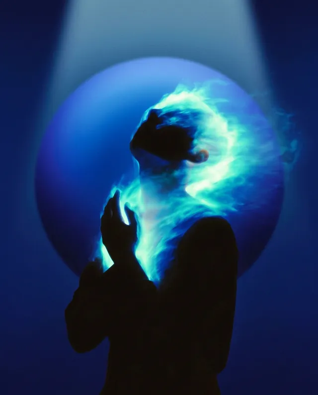 A silhouette of a figure, illuminated by ethereal blue flames, stands in a prayer-like pose against a deep blue background, creating a mystical and contemplative mood. The image uses a blend of digital art and photography, emphasizing vibrant blues and contrasts with darker tones.