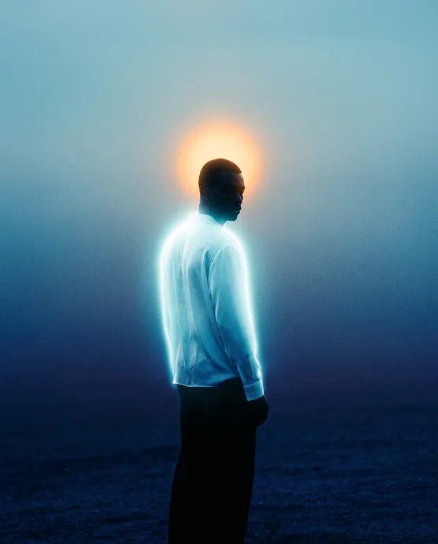 A silhouette of a man stands against a hazy backdrop, with a luminous halo effect created by the sun rising behind him. The image features a dreamy, ethereal style with a blend of cool blues and warm sunlit tones, evoking a sense of tranquility and introspection.