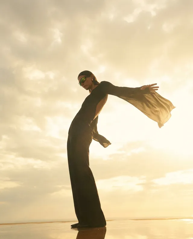 A striking editorial photograph captures a model in a dramatic black outfit, striking a dynamic pose against a backdrop of a golden sunset. The image evokes a sense of freedom and sophistication, highlighted by the warm tones of the sky and deep shadows.