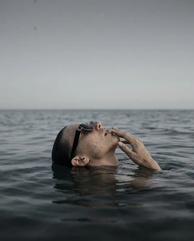 A serene editorial photograph captures a contemplative individual floating in calm waters, with subtle glitter on their skin reflecting dim light. The color palette features muted blues and grays, evoking a tranquil and introspective mood.