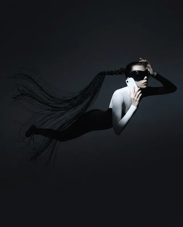 A striking editorial photograph features a figure in a monochrome bodysuit, gracefully suspended underwater while holding a phone, evoking a sense of mystery and modernity. The mood is surreal and ethereal, dominated by deep blacks and stark whites, accentuated by the flowing hair trailing behind.
