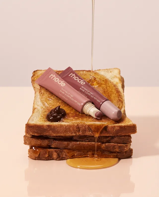 This editorial photography features a playful composition of makeup products placed on a stack of toasted bread, with honey dripping from above. The warm, soft color palette creates a lighthearted and inviting mood.