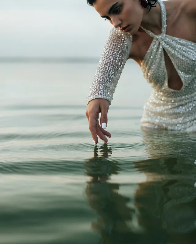 A model in a shimmering, beaded white dress reaches into calm water, capturing a serene and ethereal mood. The soft, muted colors of the scene create a tranquil atmosphere, highlighting the delicate interplay between the fabric and the water.