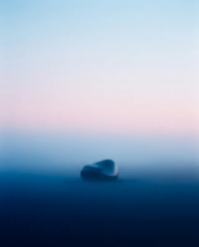 A minimalist photograph featuring a solitary abstract shape against a softly blurred gradient of pastel blues and pinks, evoking a serene and tranquil mood. The ethereal quality of the image emphasizes the simplicity and isolation of the main subject.