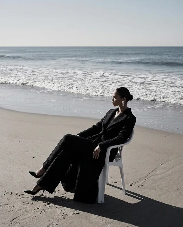 A serene editorial photograph captures a woman in a black outfit seated on a plastic chair by the ocean. The mood is calm and contemplative, with soft natural light accentuating the muted tones of the beach and water.