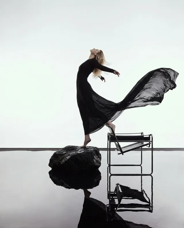 An editorial fashion photograph featuring a poised model in a flowing black dress, artfully posed on a rock with a minimalist backdrop. The mood is dramatic and ethereal, enhanced by the stark contrast of black and white tones.