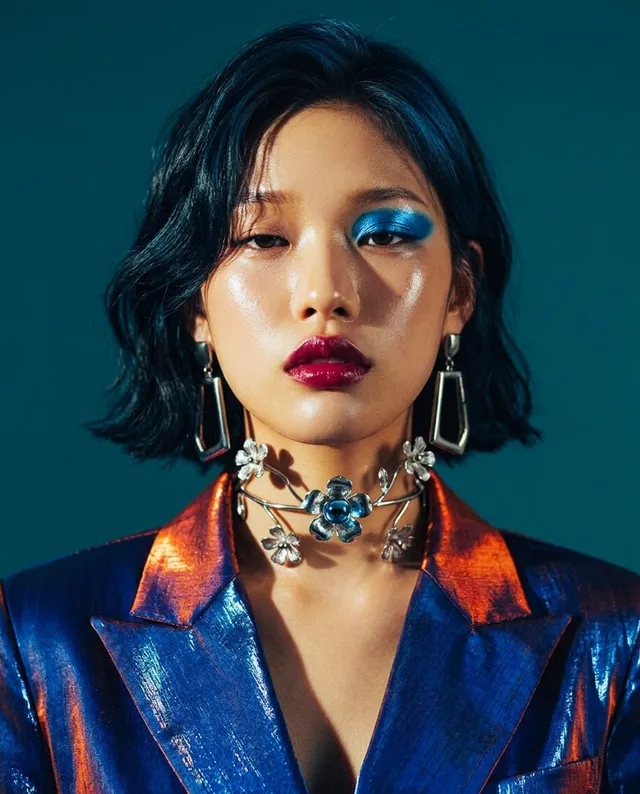 A striking editorial portrait featuring a model with a modern bob hairstyle and bold makeup, including blue eyeshadow and deep red lips. The vibrant colors of her metallic blazer—shimmery blue and orange—contrast against a dark teal background, creating a dramatic and glamorous mood.