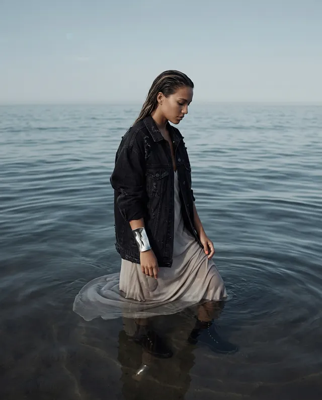 A model stands in shallow water, dressed in a flowing beige dress layered with a black denim jacket, creating a striking contrast against the soft blues of the ocean. The editorial photography captures a serene and contemplative mood, enhanced by the muted color palette.