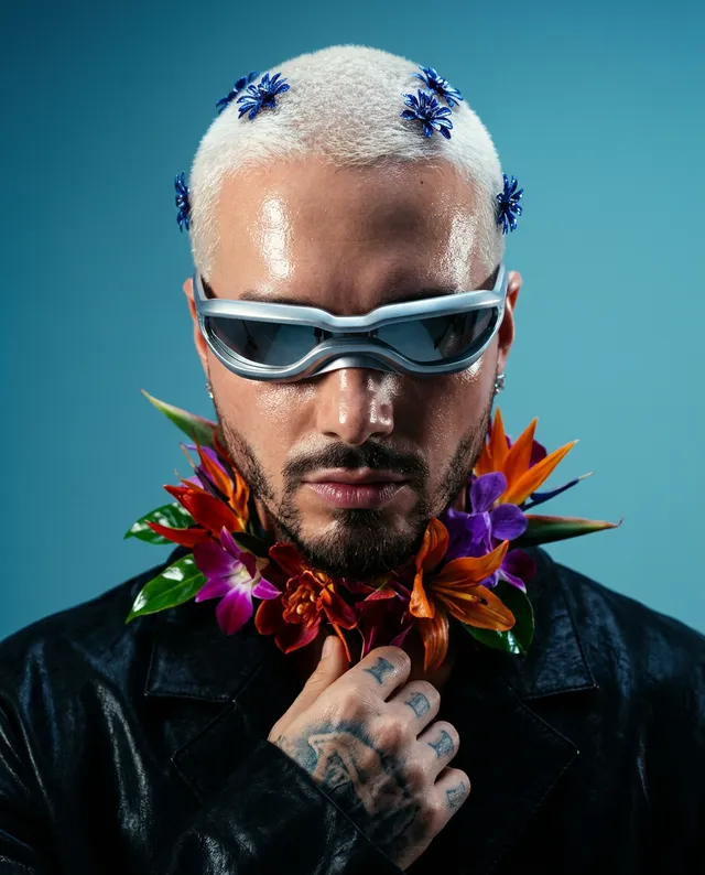 A striking portrait features a man with a buzz cut adorned with blue flowers, wearing sleek sunglasses and a vibrant floral necklace, set against a soft blue backdrop. The image, styled in editorial photography, conveys a bold and edgy mood through its vivid colors and contemporary aesthetic.