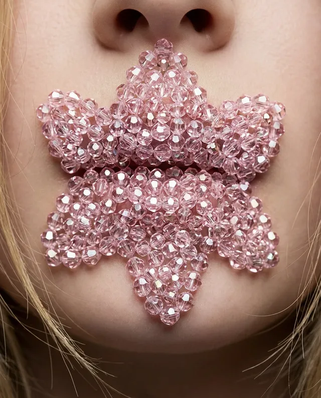 A close-up editorial photograph features a child's face adorned with a large, intricate pink beaded flower over their mouth, creating a surreal and artistic effect. The dominant colors are soft pinks and neutral skin tones, evoking a whimsical and playful mood.