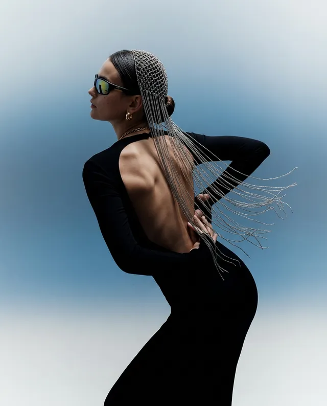 A striking editorial photograph features a model in a sleek black dress, showcasing an open back and adorned with a unique headpiece. The mood is contemporary and fashion-forward, with a gradient blue background enhancing the dramatic silhouette and reflective sunglasses.