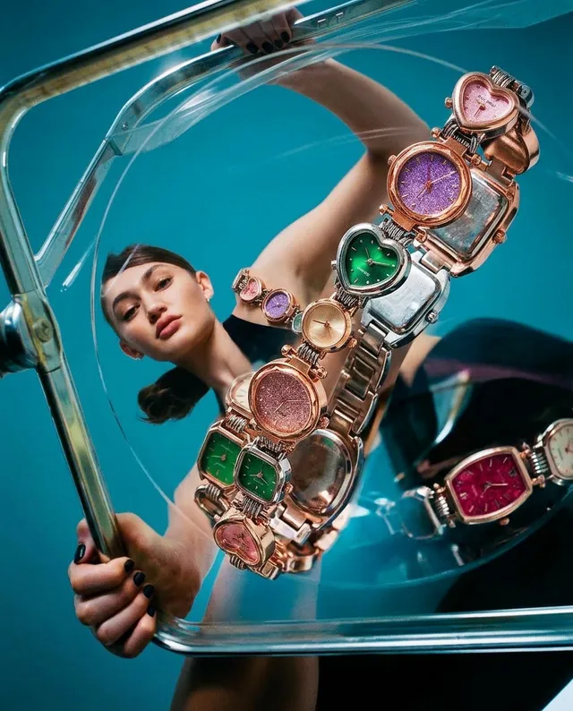 A striking editorial photograph featuring a model gazing through a transparent display case that holds an array of colorful, stylish watches. The vibrant color palette includes greens, pinks, and purples against a cool blue background, creating a modern and playful mood.