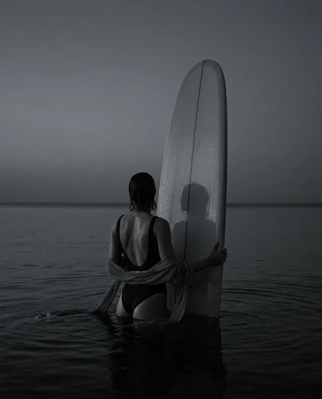 A monochromatic editorial photograph captures a lone figure in a swimsuit standing in calm water, holding a surfboard while gazing toward the horizon. The mood is tranquil and introspective, with dominant shades of gray and soft lighting enhancing the serene atmosphere.