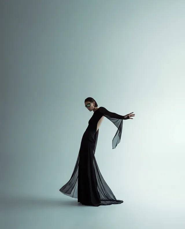 A striking editorial photograph features a model in a flowing black gown with sheer sleeves, poised dramatically against a soft, light backdrop. The mood is avant-garde and sophisticated, highlighted by the contrast of deep black and soft pastel tones.