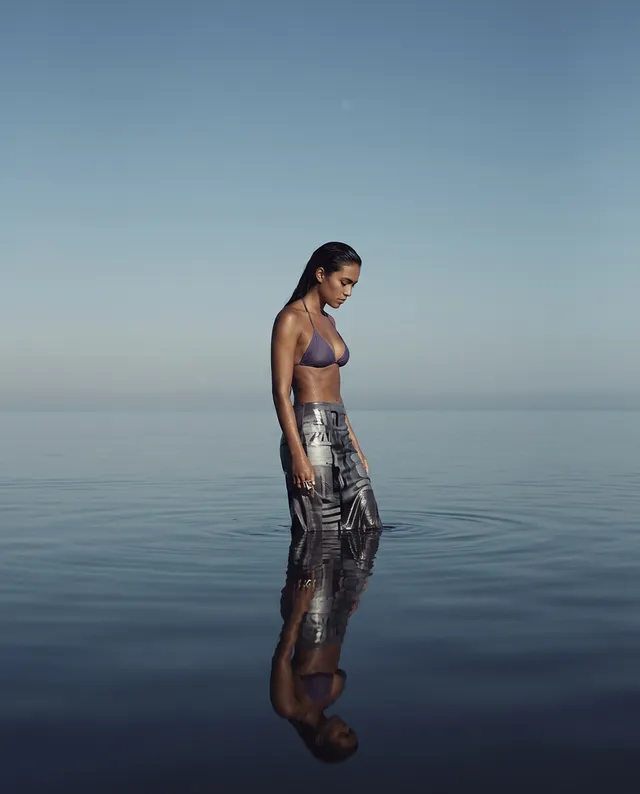 A serene editorial photograph features a woman standing in calm waters, reflecting her tranquil demeanor against a soft blue sky. The dominant colors are cool tones of blue and gray, evoking a peaceful and contemplative mood.