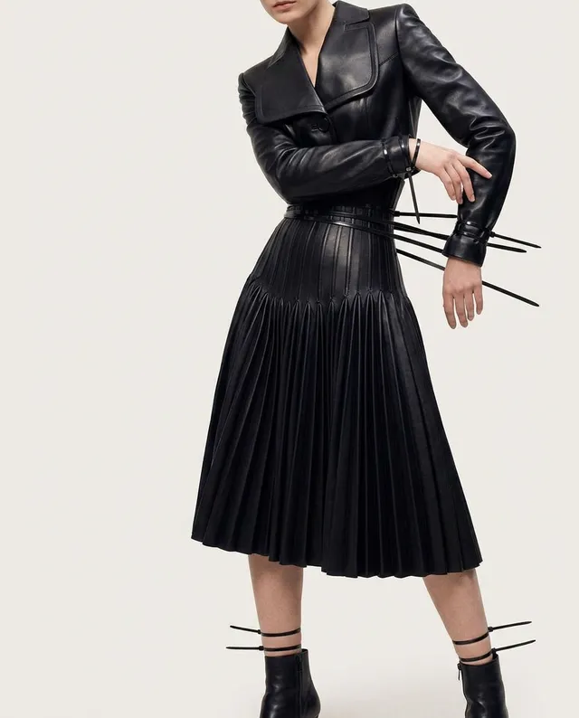 This editorial fashion photograph features a model in a sleek black leather dress with pleated details, exuding a sophisticated and powerful mood. The dominant colors are shades of black, set against a soft, neutral background that highlights the garment's texture and design.