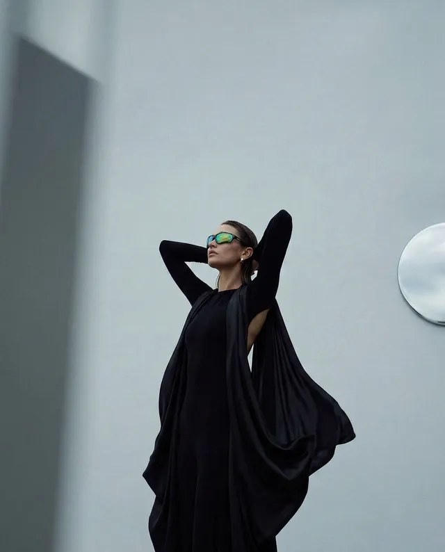 A striking editorial photograph features a model in a flowing black gown with elongated sleeves, set against a minimalist gray backdrop. The mood is bold and dramatic, accentuated by her confident pose and the reflective sunglasses, creating a modern and sophisticated aesthetic.