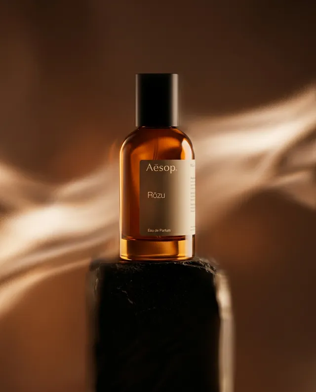 A sleek glass bottle of Aesop's Rōzu perfume stands elegantly atop a dark pedestal, exuding a sense of luxury. The warm amber tones and soft, blurred background create a soothing, sophisticated atmosphere.