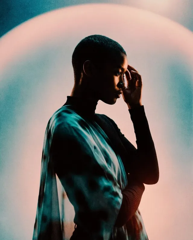 A striking editorial portrait features a silhouette of a woman with a shaved head, elegantly draped in a translucent fabric, set against a soft, luminous halo of pastel colors. The mood is contemplative and ethereal, enhanced by the interplay of shadows and light.