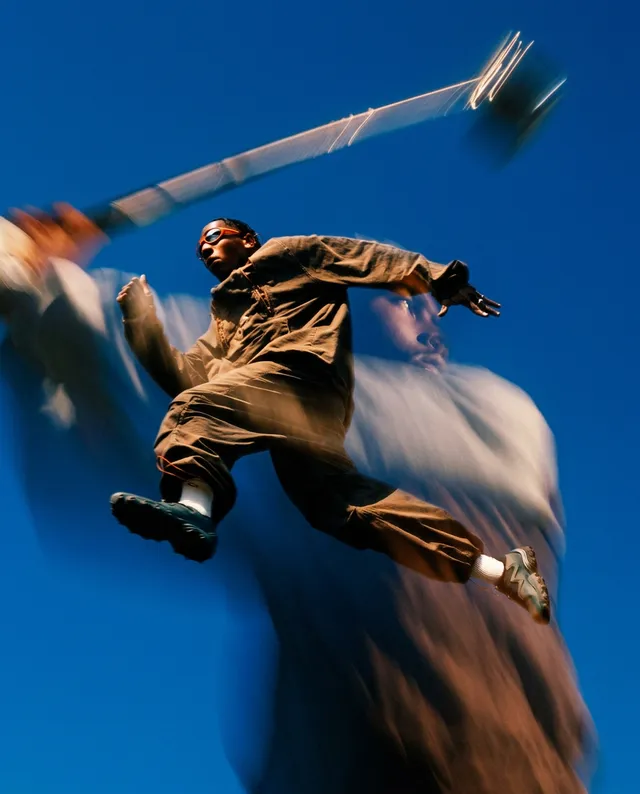 A dynamic editorial photograph captures a figure leaping with a sense of energy and motion against a vibrant blue sky. The dominant colors are deep blues and earthy tones, evoking a lively and exhilarating mood through a blurred effect that enhances the sense of action.