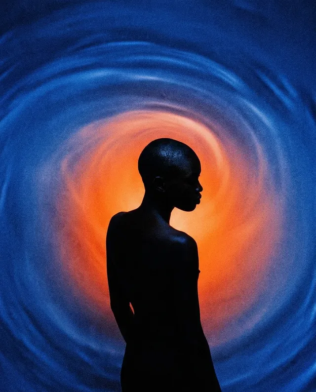 A striking silhouette of a person with a shaved head contrasts against a swirling backdrop of deep blue and vibrant orange hues. The editorial photography evokes a sense of mystery and introspection.