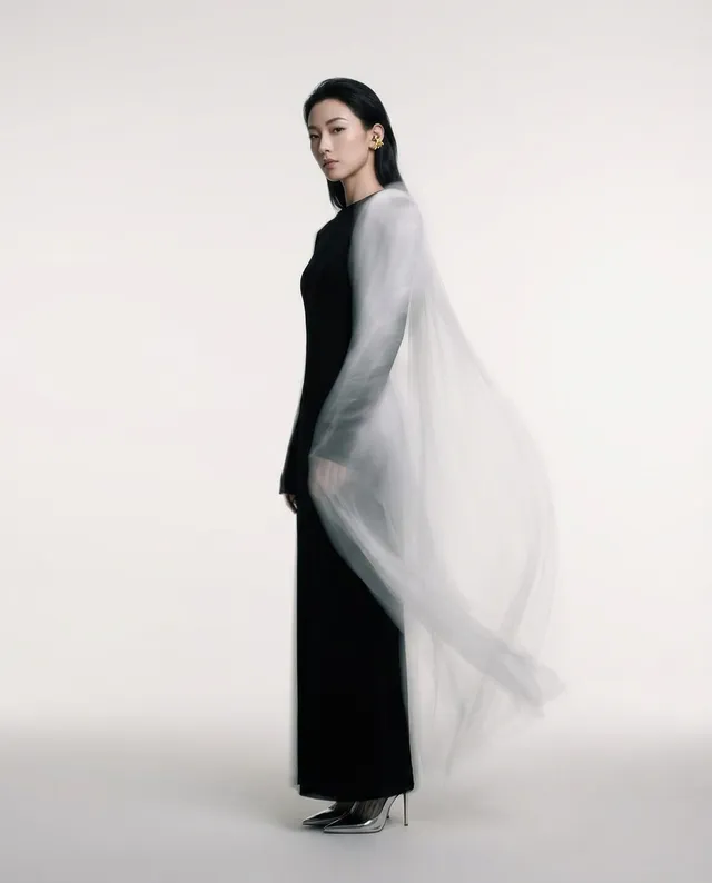 A striking editorial photograph featuring a poised woman in a sleek black gown, accentuated by a flowing sheer cape. The minimalistic background and soft, ethereal mood highlight the elegance of the subject, with dominant shades of black and light grey.