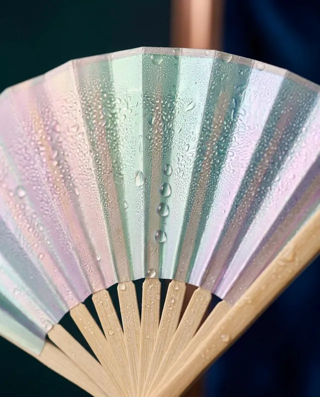 This close-up editorial photograph features an iridescent fan adorned with droplets of water, capturing a fresh and vibrant mood. The dominant colors are soft pastel hues, creating a serene and elegant aesthetic against a dark background.