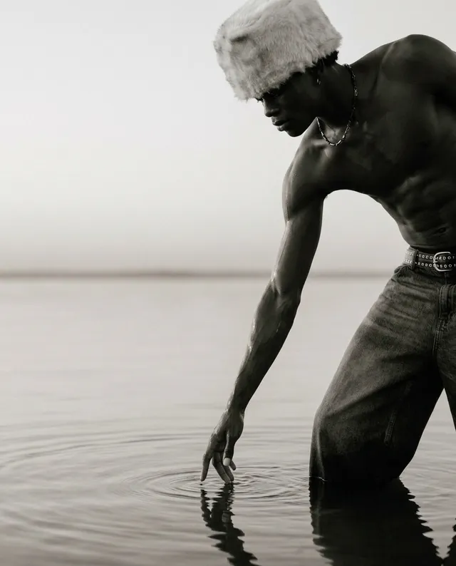This black-and-white editorial photograph features a man gently reaching into the water, creating ripples. The mood is serene, accentuated by the soft fading light and natural surroundings.