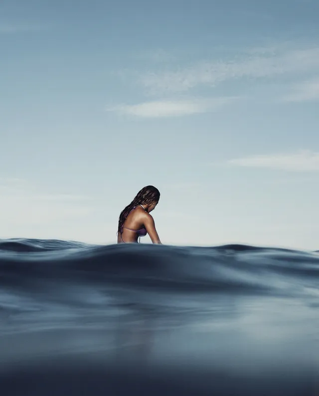 A serene editorial photograph captures a solitary figure submerged in calm ocean waters, evoking a sense of introspection and tranquility. The dominant blues and soft grays create a peaceful, soothing mood against the vast sky.