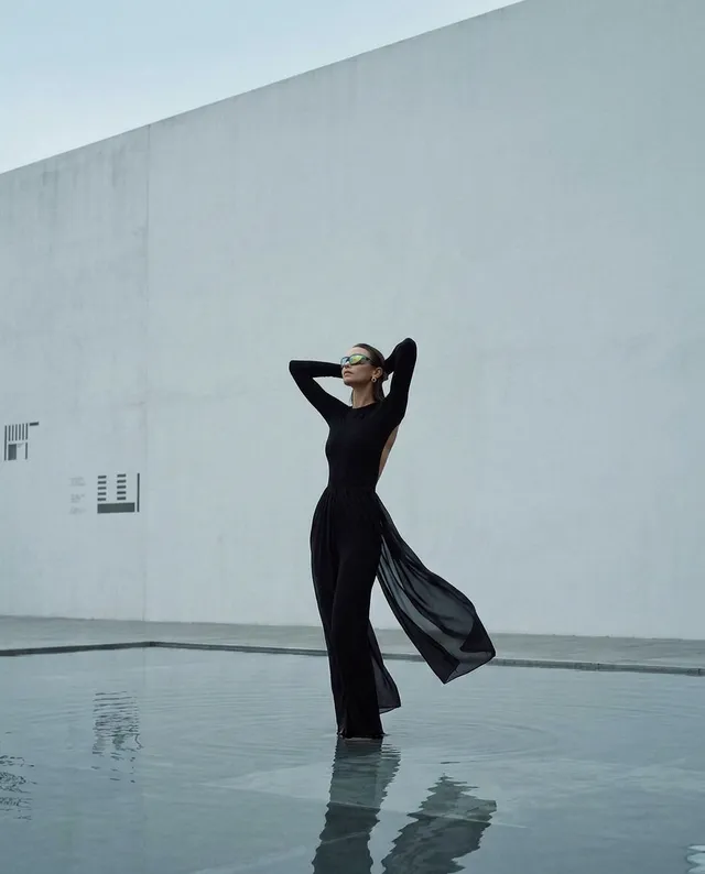 A striking editorial photograph features a model in a flowing black outfit standing in shallow water, with a minimalist white wall in the background. The serene mood is enhanced by cool tones and reflections, creating a sense of tranquility and elegance.